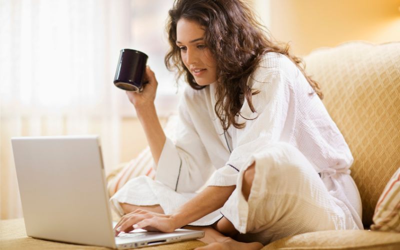 Woman with coffee in hand and working on laptop on bed